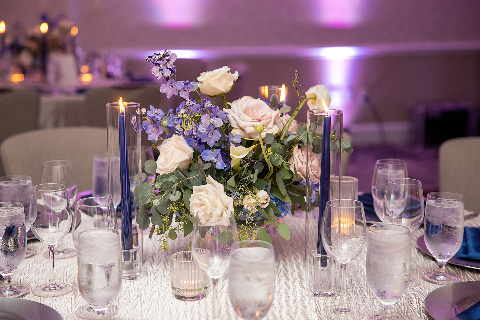 A glass compote vase with white and blue flowers, surrounded by navy blue taper candles.