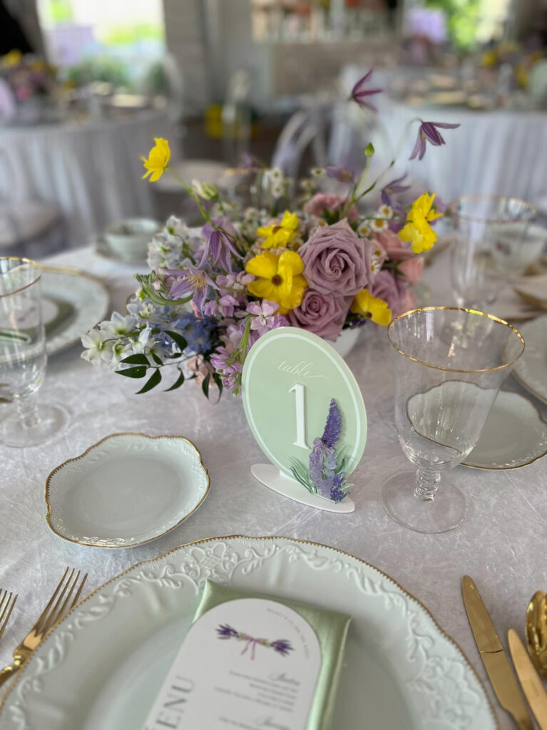 A white vase with purple, pink and yellow pastel flowers on a table surrounded by glassware and white plates with gold rim